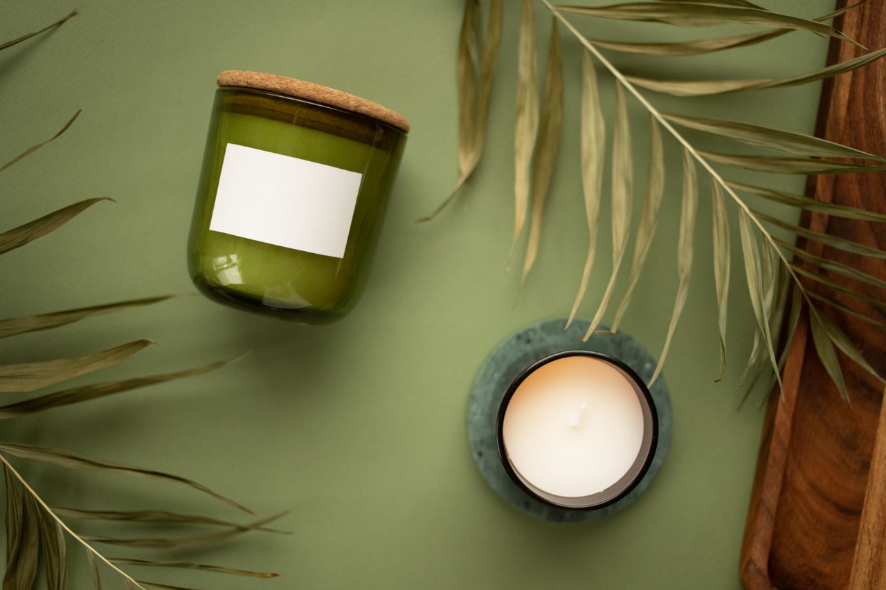 our-journey-03 Aesthetic display of a green jar candle and a lit scented candle with natural leaves on a green background.